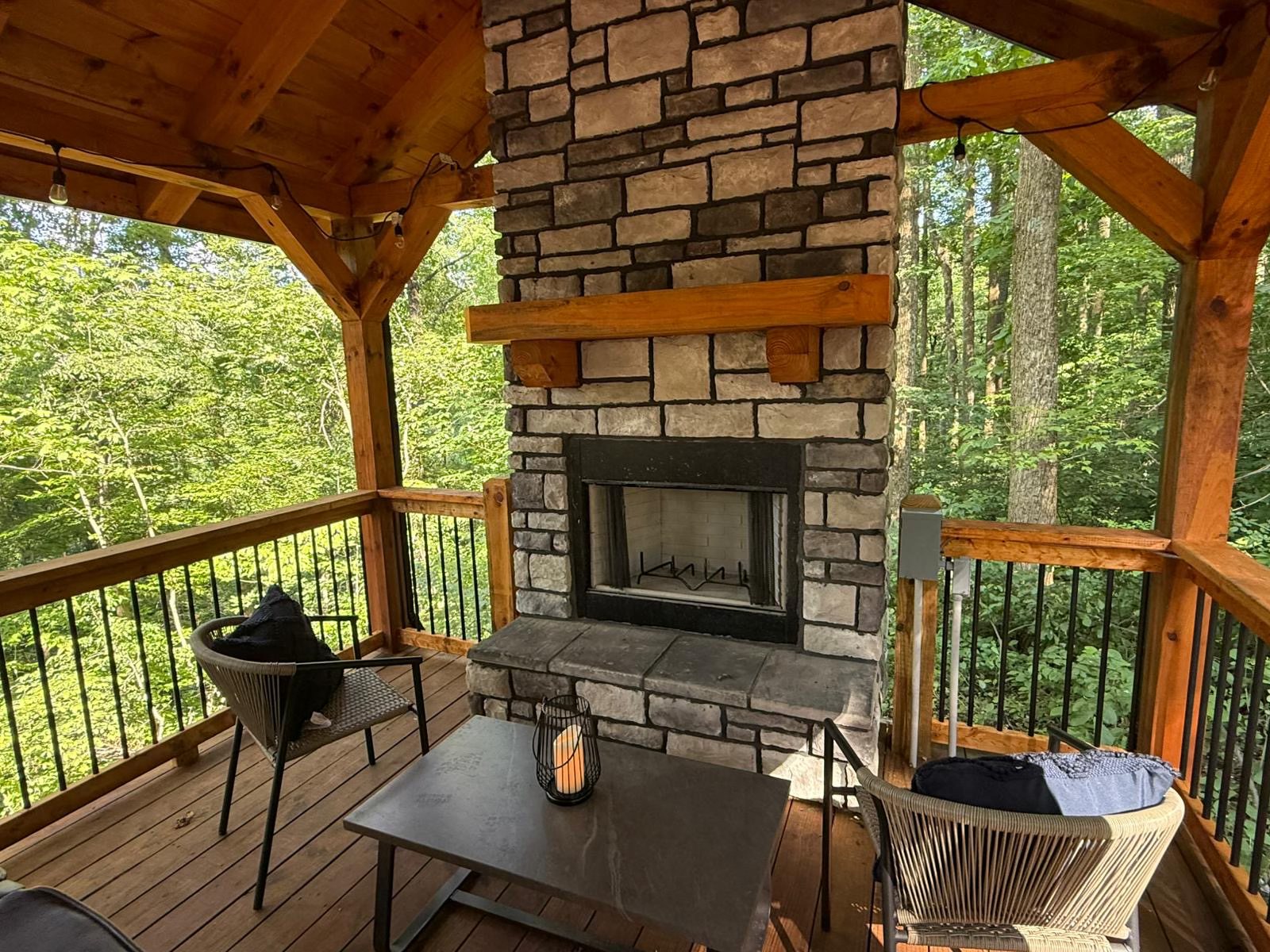 A cozy wooden deck with a stone fireplace is surrounded by lush green forest. Two chairs and a table with a candle create a serene, inviting atmosphere.