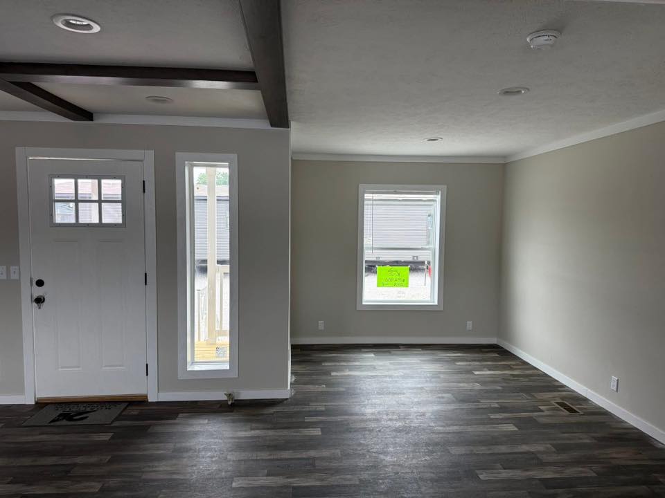 Modern, unfurnished room with grayish wood floor and light-colored walls. Features a white front door with windows, and another large window displaying an outside view.