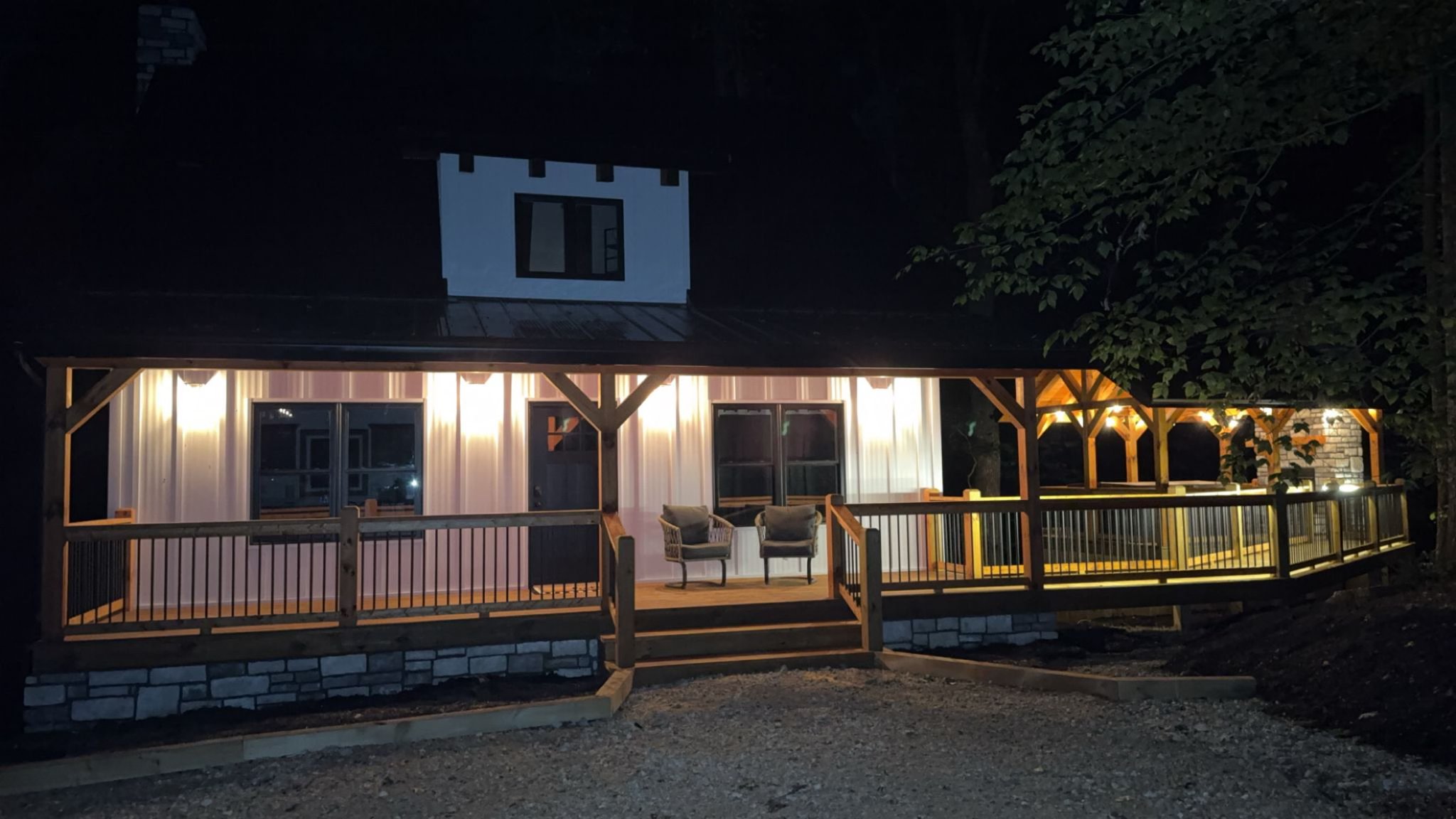 A cozy cabin porch at night, warmly lit with soft lights. Wooden railings and chairs create an inviting ambiance, surrounded by darkness and lush trees.