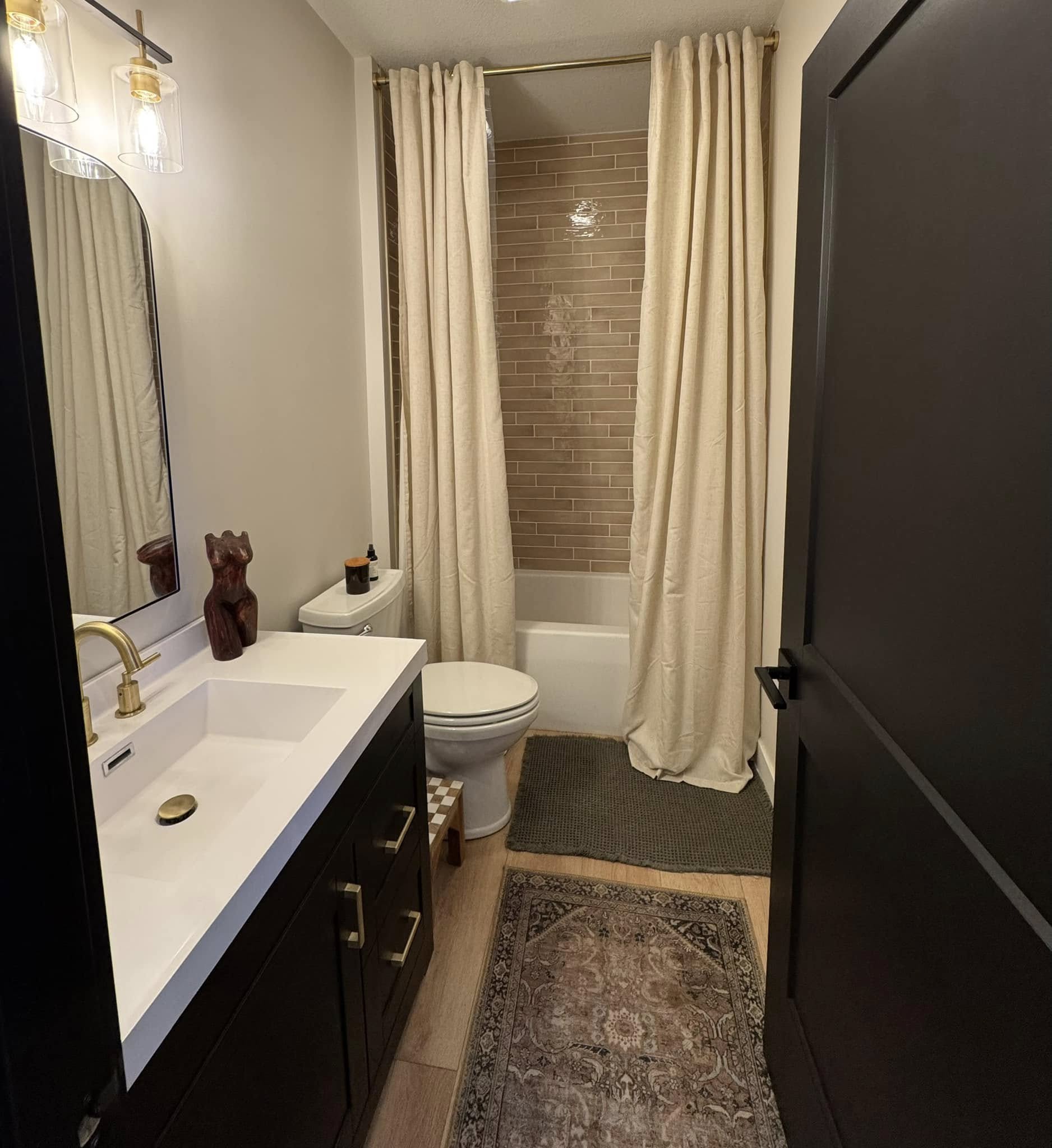 Modern bathroom with dark vanity, gold fixtures, and a white sink. Beige curtains frame a tiled bathtub. A patterned rug adds warmth on wood floors.