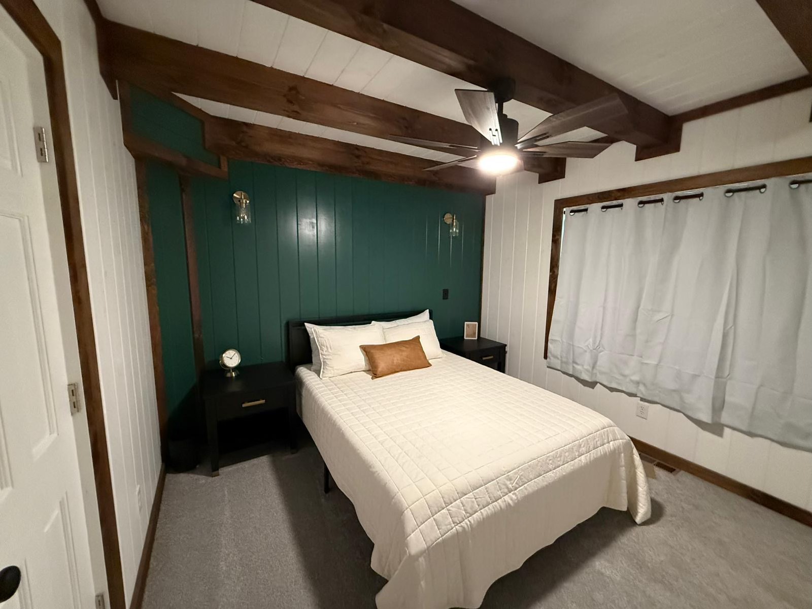 Bedroom with a cozy feel, featuring a bed with white and tan pillows. Green accent wall, wooden beams, ceiling fan, side tables, and curtained window.