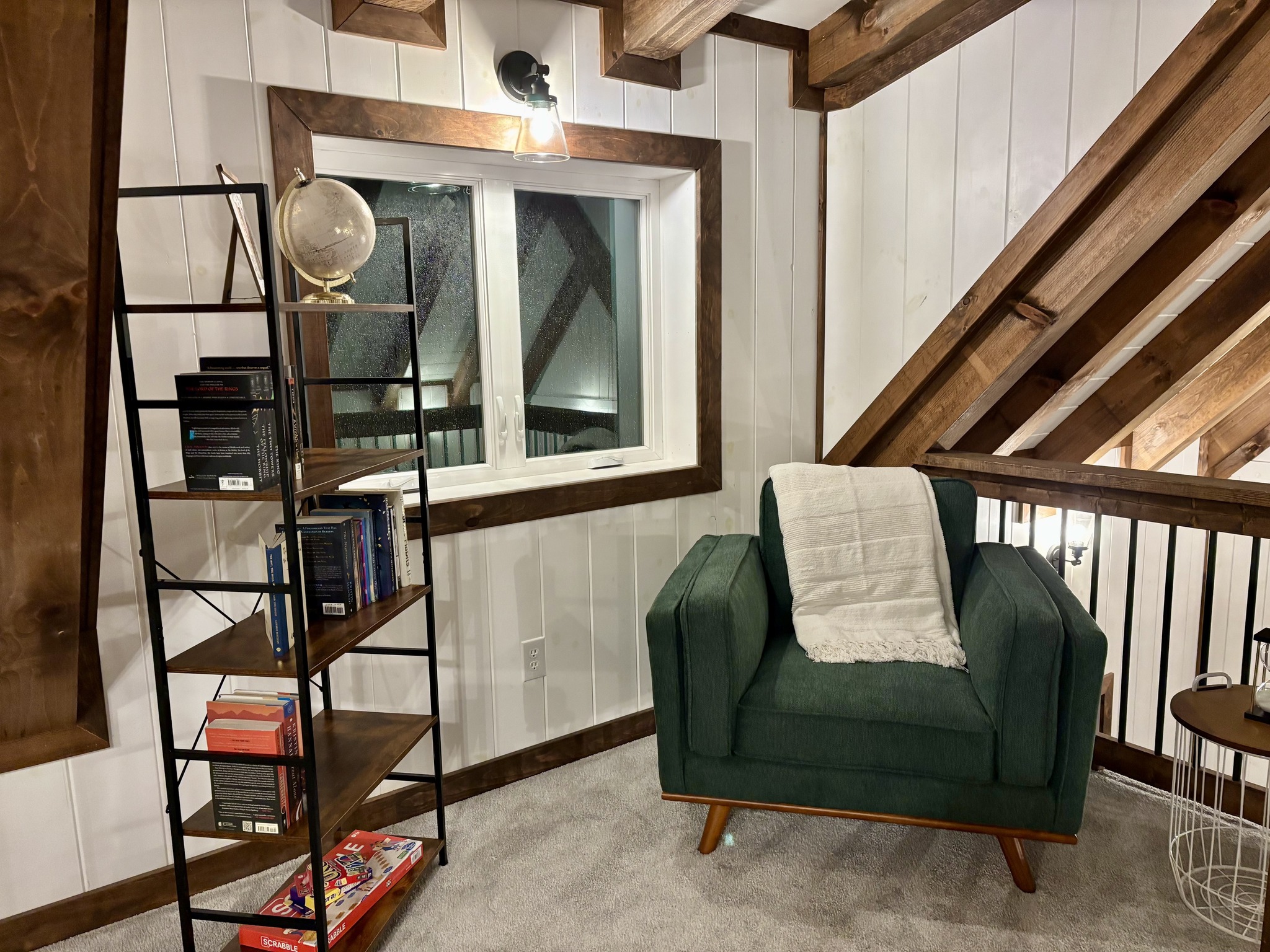 Cozy reading nook with a green armchair draped with a white throw, under warm lighting. Nearby, a wooden shelf holds books and a globe, enhancing a rustic vibe.