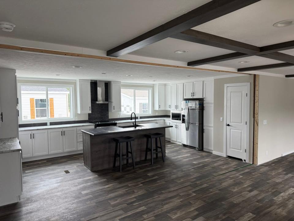 Modern kitchen with dark wood flooring, white cabinets, and a gray island with bar stools. Large windows provide natural light, creating a bright, open space.