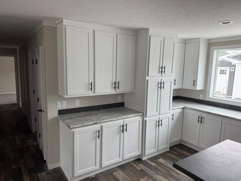 Modern kitchen with white cabinets, marble countertops, and dark handles. Bright window and wood floor create a fresh, clean ambiance.