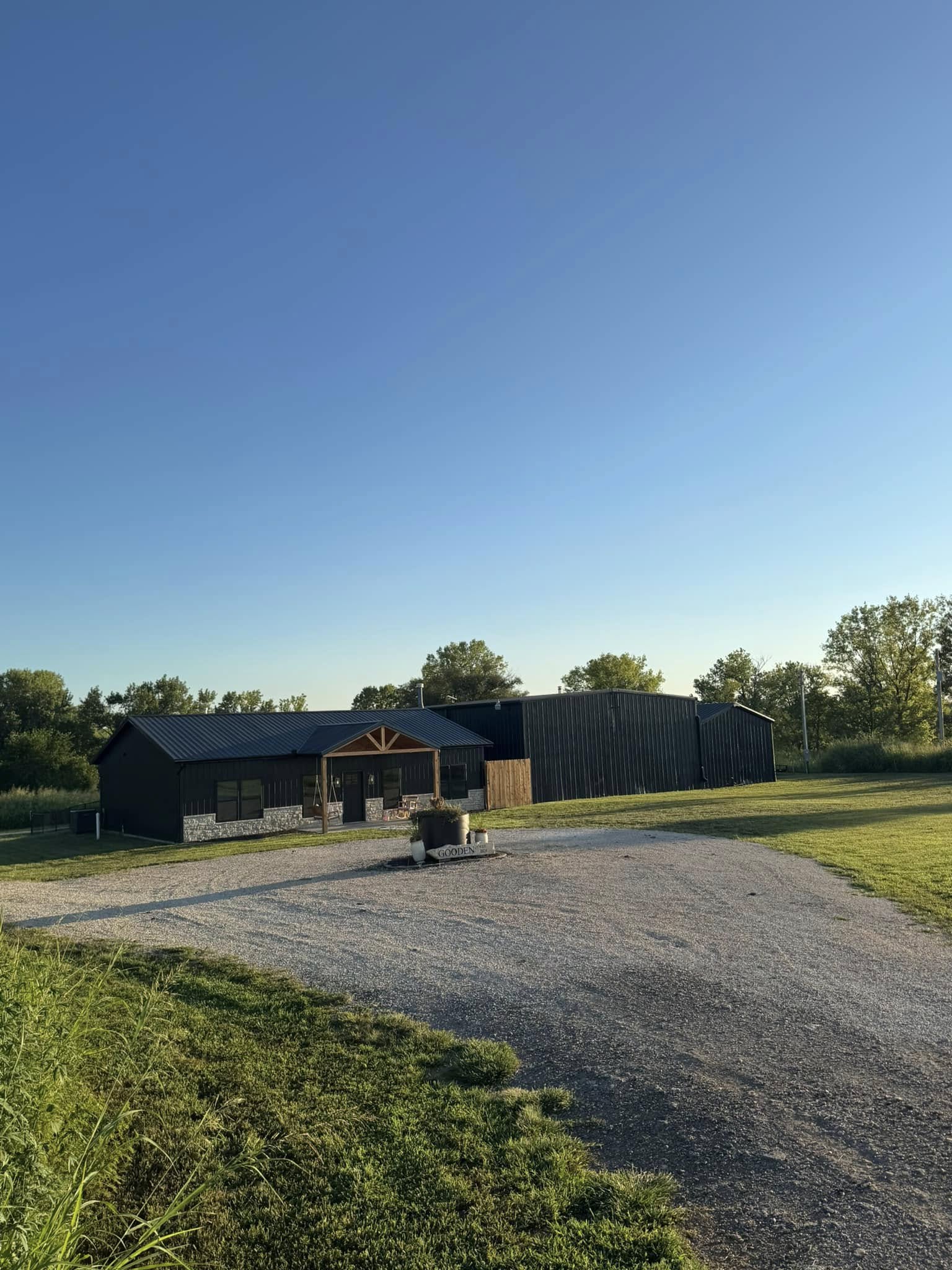 A small, dark-colored barn with a wooden porch sits in a sunny, rural area. Gravel driveway and green grass surround it, with trees in the background.