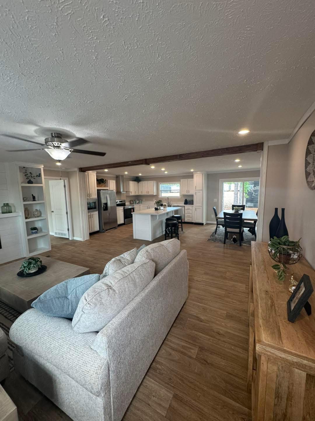 Modern open-plan living space with neutral tones. Features a light gray couch, hardwood floor, ceiling fan, spacious kitchen, and a dining area. Cozy and inviting atmosphere.