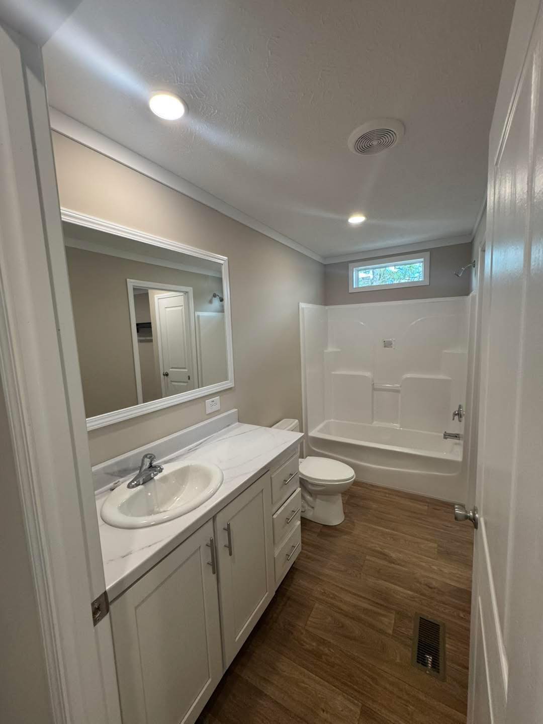 Modern bathroom with white fixtures, large mirror, wood-look tile floor, and a shower-tub combo. Soft lighting creates a clean, inviting ambiance.