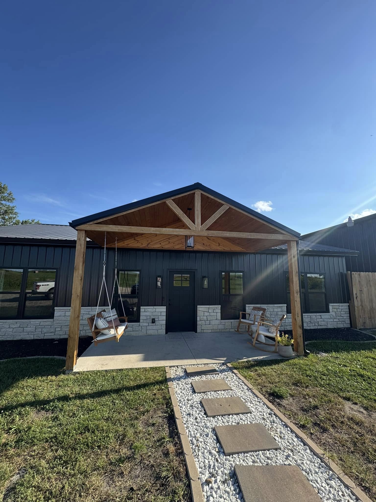 A modern house with a wooden porch features a swing and bench. A stone path leads to the door. The blue sky adds a serene, inviting atmosphere.