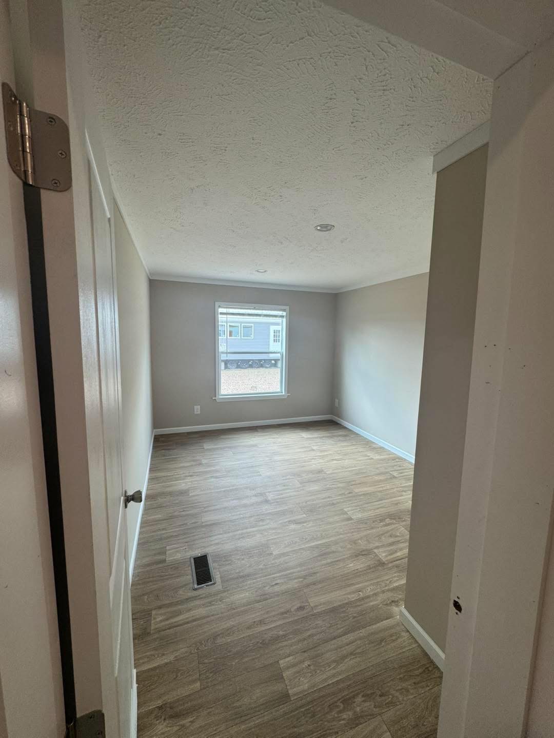 Empty room with wooden floors, beige walls, and a white ceiling. A single window lets in natural light, creating a calm and minimalist atmosphere.