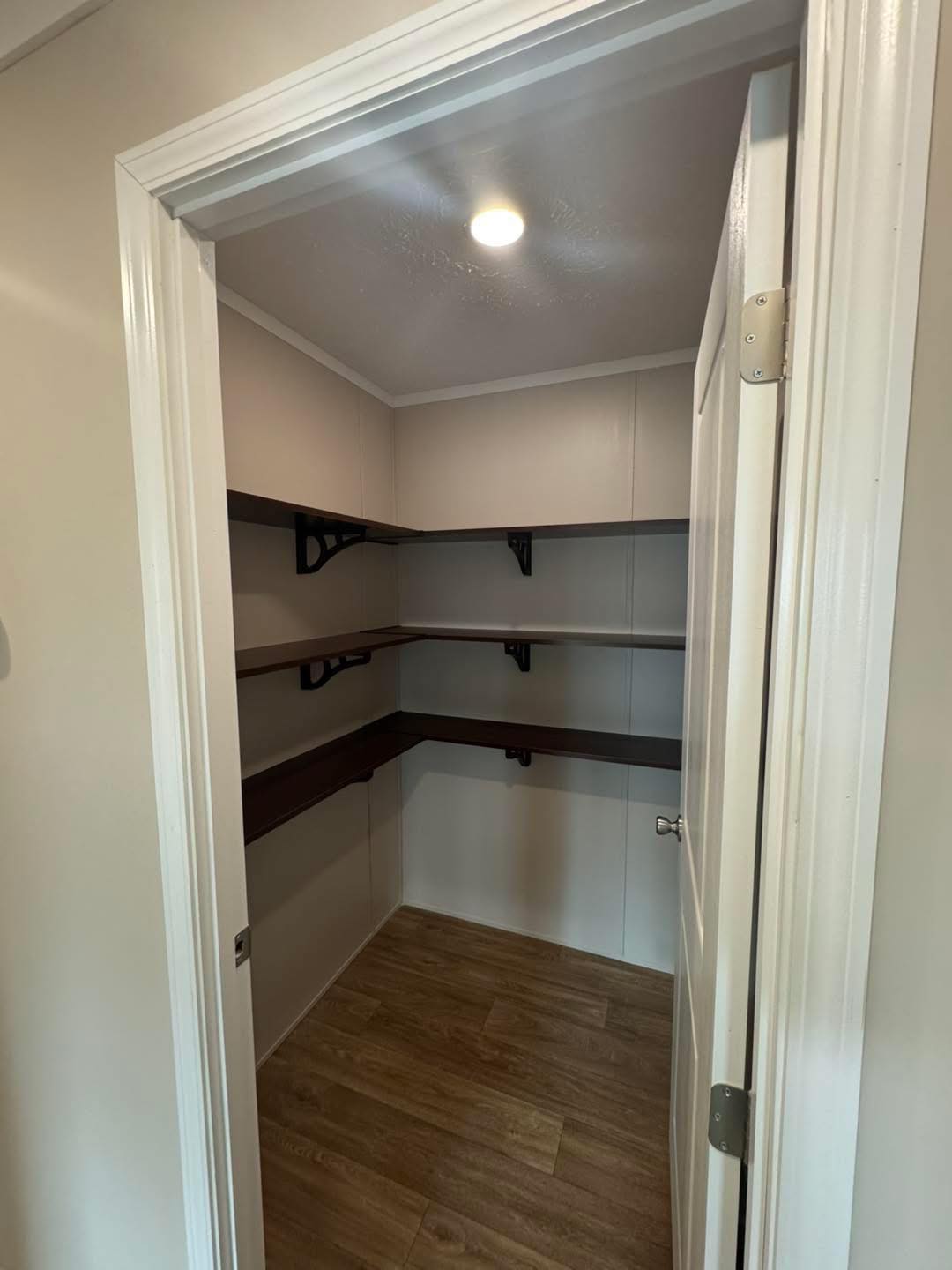 Small empty closet with wooden shelves on three walls, illuminated by a ceiling light. The floor is wood-patterned, creating a clean and functional space.