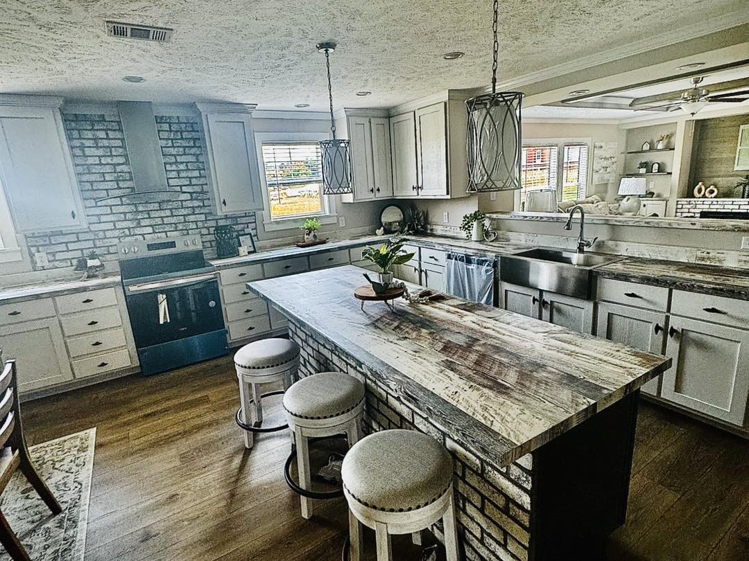 Spacious kitchen with rustic charm, featuring a large stone island, three cushioned stools, brick backsplash, stainless steel appliances, and pendant lights. Cozy and inviting.