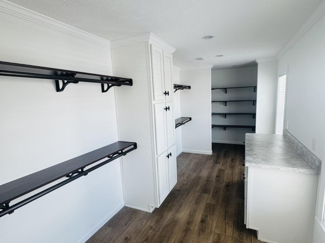 Bright white pantry with dark shelves and wood flooring. Cabinets with black handles line one wall, while open shelving adorns the other, creating a modern, organized feel.