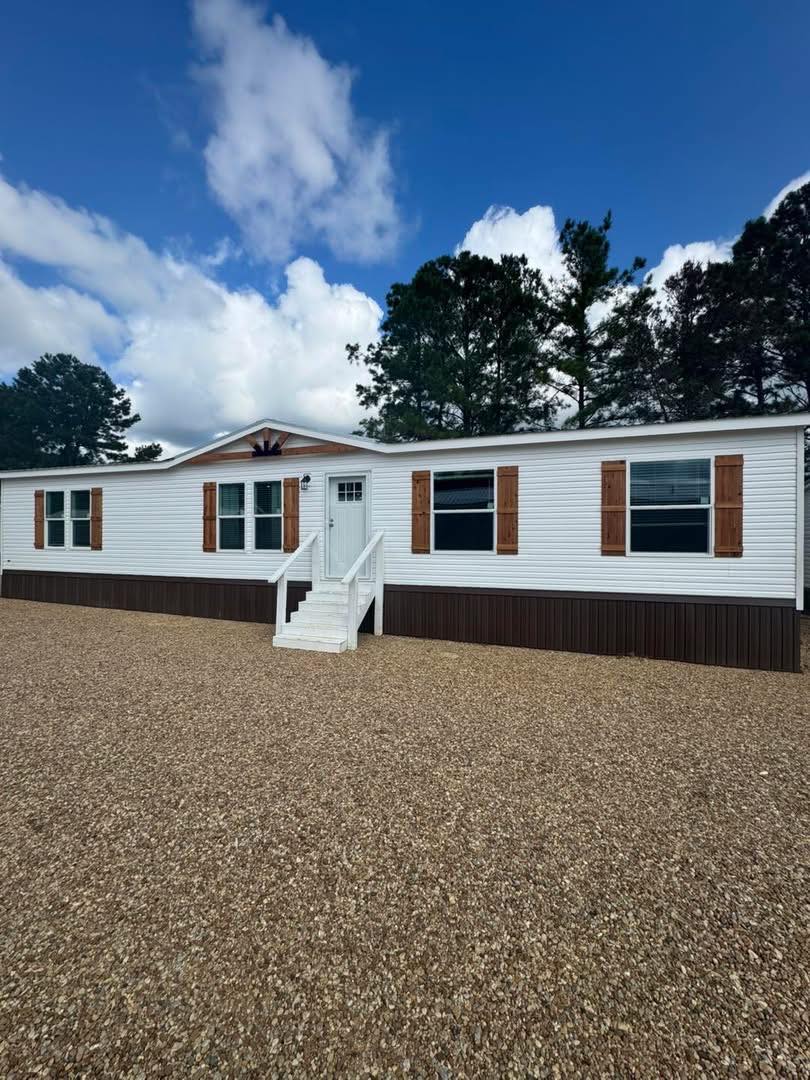 A white manufactured home with a brown base and wooden shutters stands on a gravel lot. Tall trees and a blue sky with fluffy clouds are in the background.