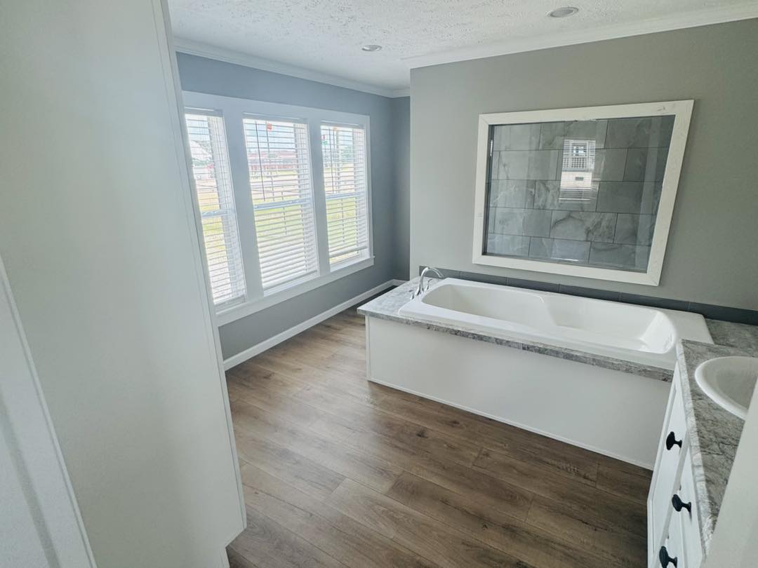 Bright bathroom with wood flooring, featuring a white standalone bathtub and a marble countertop. Three large windows let in natural light, creating a serene atmosphere.