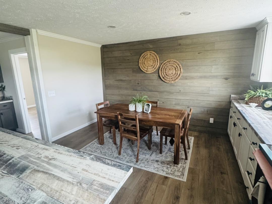 Cozy dining area with a rustic wooden table, six chairs, and potted plants. Wooden accent wall with two woven decorations conveys a warm, inviting atmosphere.