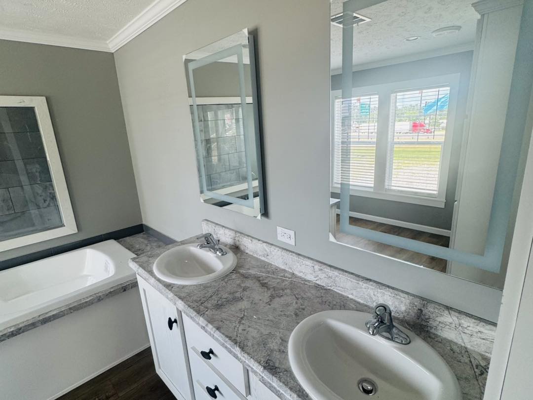 Modern bathroom with dual sinks and mirrors, gray marble countertop, and a large bathtub. Sunlight streams through a window, creating a bright, clean atmosphere.