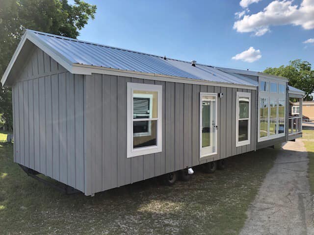 Gray tiny house with large windows and a metal roof, situated on a grassy area under a blue sky with fluffy clouds, conveying a cozy and modern vibe.