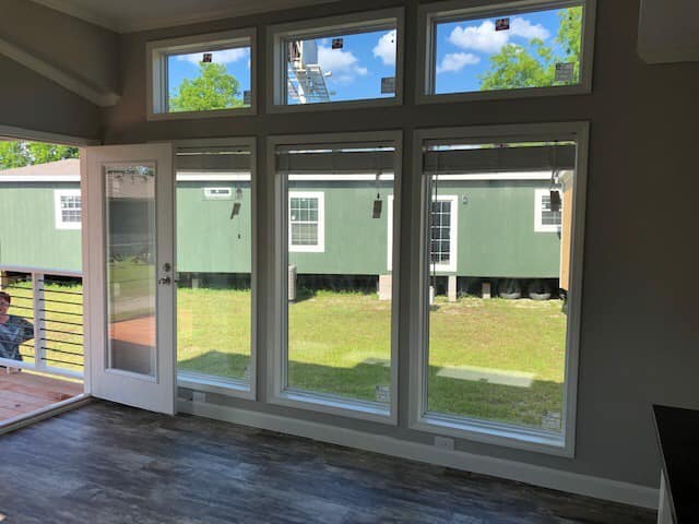 Spacious room with dark wooden floors and large windows, revealing a bright outdoor view of green grass and a green building. Open glass door on the left.