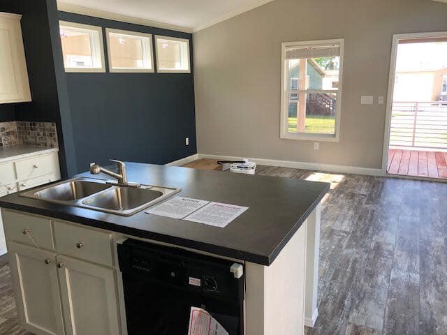 A modern, empty living space featuring a kitchen with a dark countertop and stainless steel sink. Large windows and a door lead to a sunlit patio.