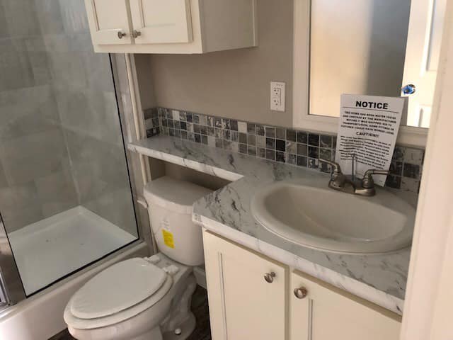 Compact bathroom with white cabinets, marble counter, and gray-tiled backsplash. Includes a shower, toilet, and sink with a "Notice" sign above. Modern and tidy.