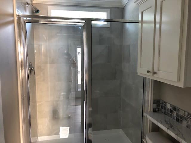 A modern bathroom with a glass shower enclosure, light gray tiles, white cabinets, and marble countertop. Soft natural light creates a calming atmosphere.