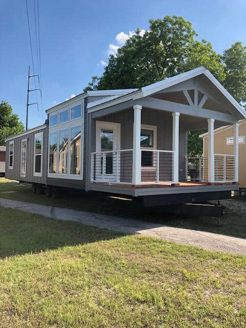 Modern tiny house with large windows and a front porch, set in a grassy area with trees and blue sky. The design is fresh and inviting.