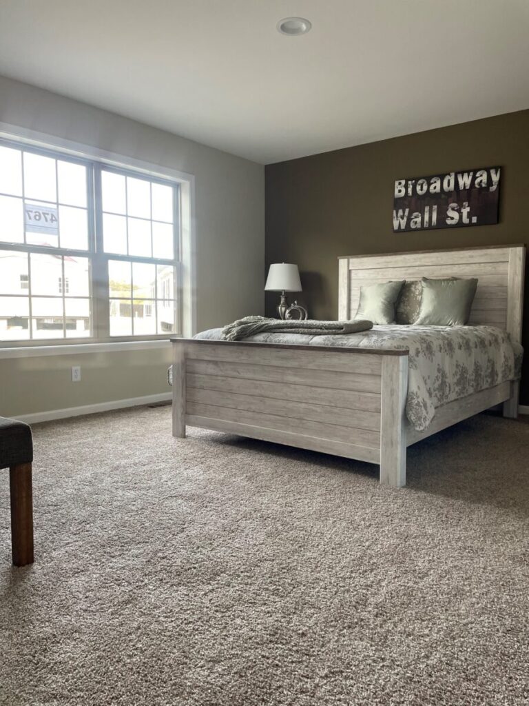 Cozy bedroom with a rustic wooden bed, floral bedding, and a lamp on the side table. Sunlight streams through a large window, illuminating a carpeted floor. A wall sign reads “Broadway Wall St.”