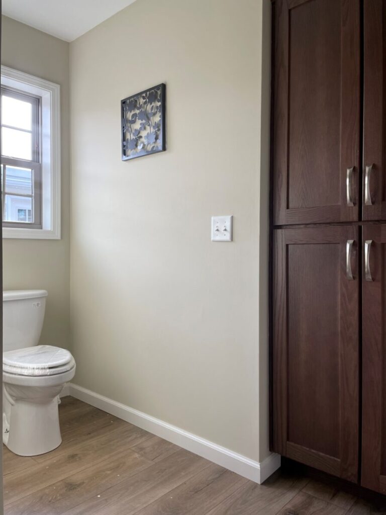 A small bathroom with light beige walls features a white toilet beside a window. A dark wooden cabinet stands on the right, with a floral picture above.