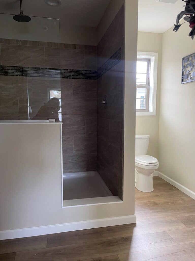 Bathroom with a modern walk-in shower featuring beige tiles and a glass partition. A toilet sits by a sunlit window, with wood flooring.