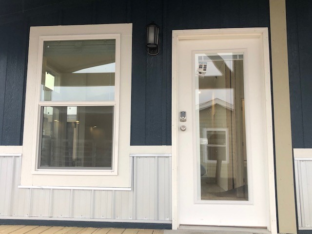Front view of a house entry. A glass door with a keypad lock and adjacent window sit within blue siding and white trim. A wall light is above.