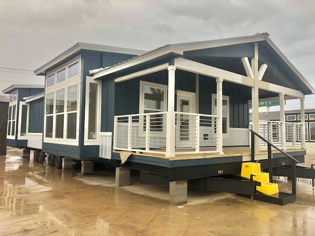Modern blue manufactured home with a porch, large windows, and white trim. The scene is overcast and the ground appears wet, suggesting recent rain.