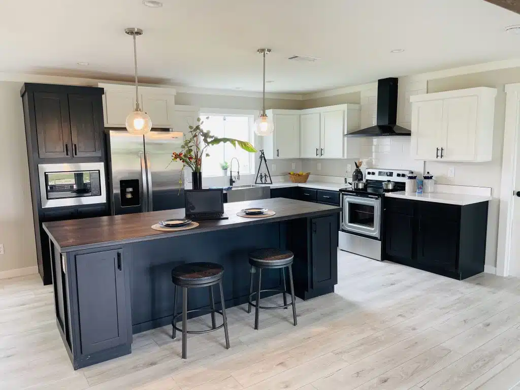 Modern kitchen with dark island, two stools, and pendant lights. Stainless steel appliances, white and black cabinets, and a vase with flowers on the counter. Bright, airy atmosphere.