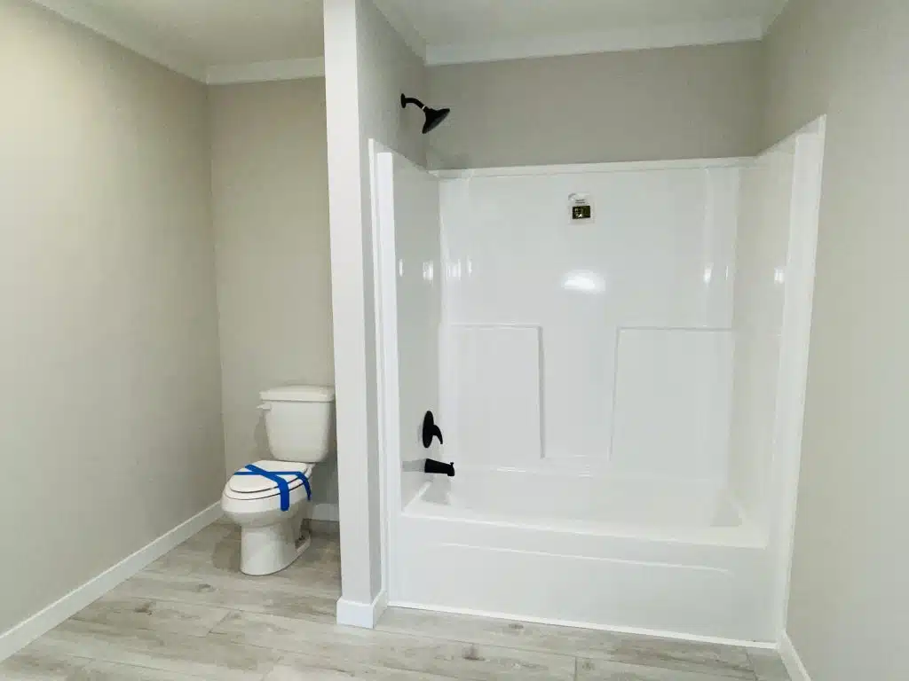 Bathroom with a white bathtub-shower combo featuring black fixtures. Adjacent is a toilet with blue tape on the lid. Light gray walls and wood floor add a clean, new look.