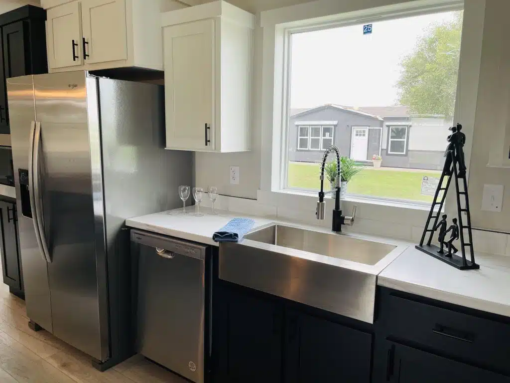 Modern kitchen with a large stainless steel fridge, sink, and dishwasher, white and black cabinets, a window view of a house, and two wine glasses.