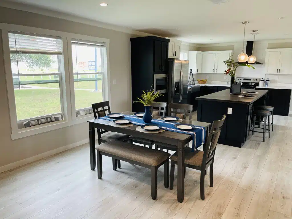 Modern dining room and kitchen with light wood flooring. A dining table with a blue runner and six plates faces an island with pendant lights. Bright and inviting.