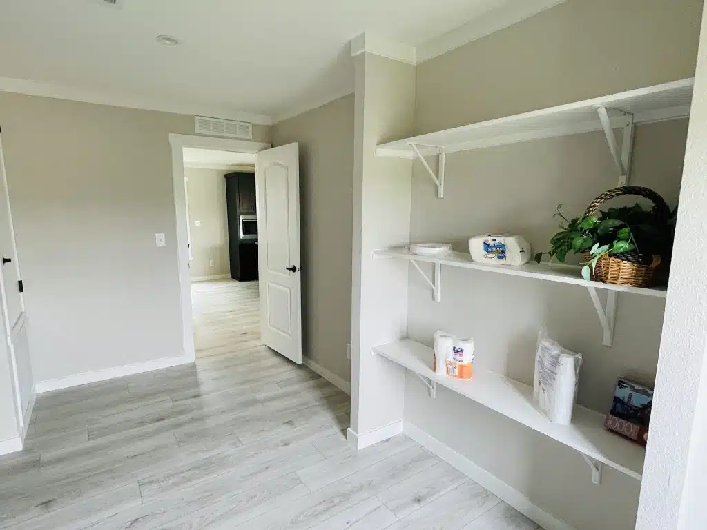A minimalist room with light gray walls and wood flooring. White shelves hold paper goods and a green plant in a basket. An open door leads to another room.