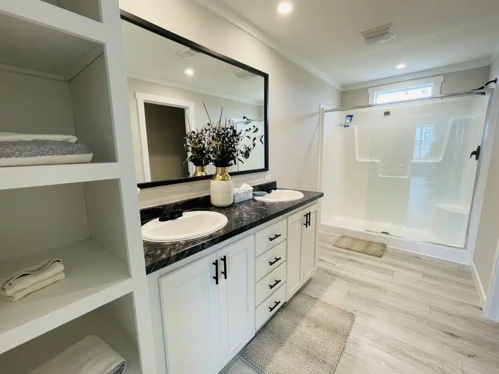 Modern bathroom featuring a double sink vanity with a black countertop and white cabinets, a large mirror, and a glass shower. Neutral tones convey elegance.