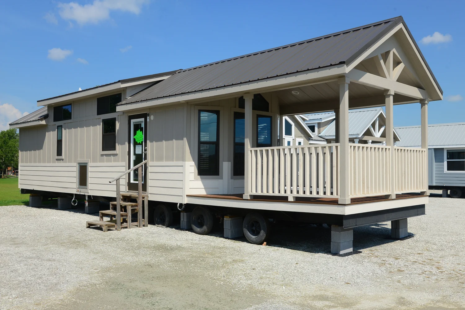 Tiny house on wheels with a gabled roof and porch, parked on gravel. It features large windows and a welcoming appearance under a blue sky.