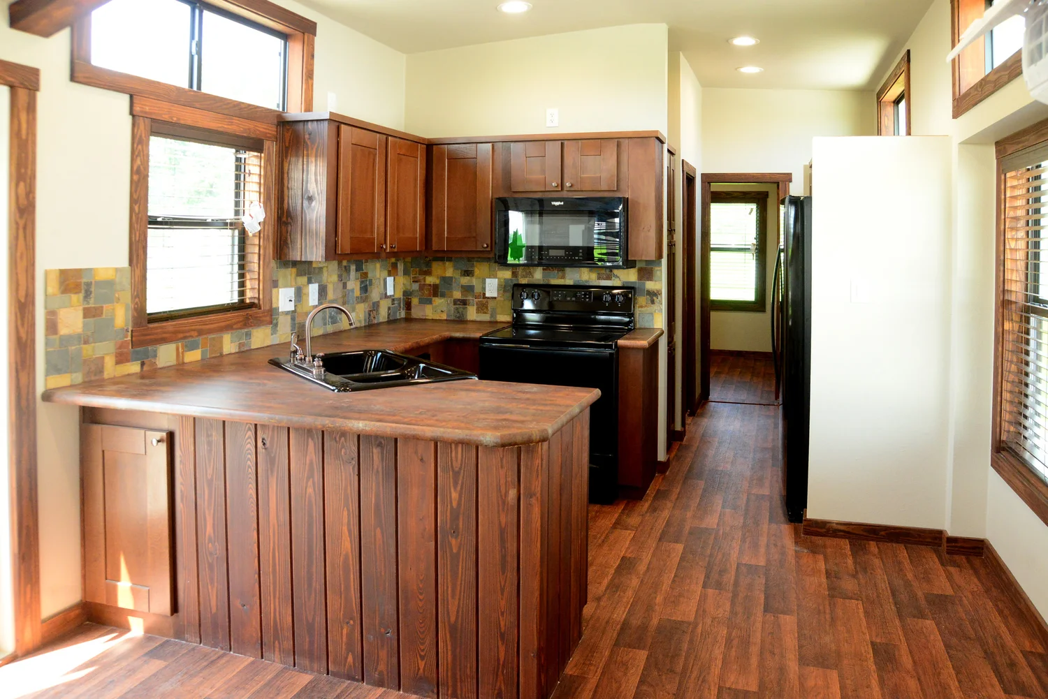 Warm-toned kitchen with wood-paneled cabinets, tiled backsplash, and dark appliances. Sunlit from large windows, creating a cozy, inviting atmosphere.