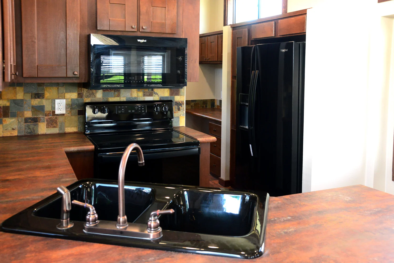 A modern kitchen with dark brown cabinets, a black sink, oven, microwave, and fridge. Warm lighting and tile backsplash create a cozy ambiance.