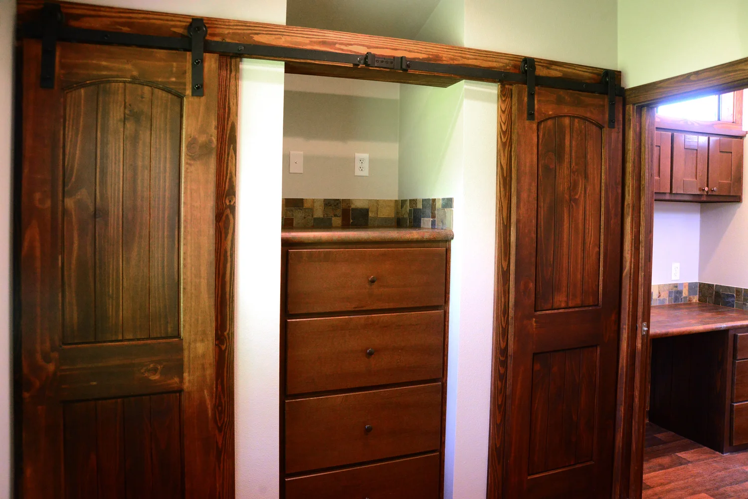 Warm-toned wooden sliding doors reveal a closet with a tall dresser inside. Rustic room ambiance with wood accents and soft natural light.