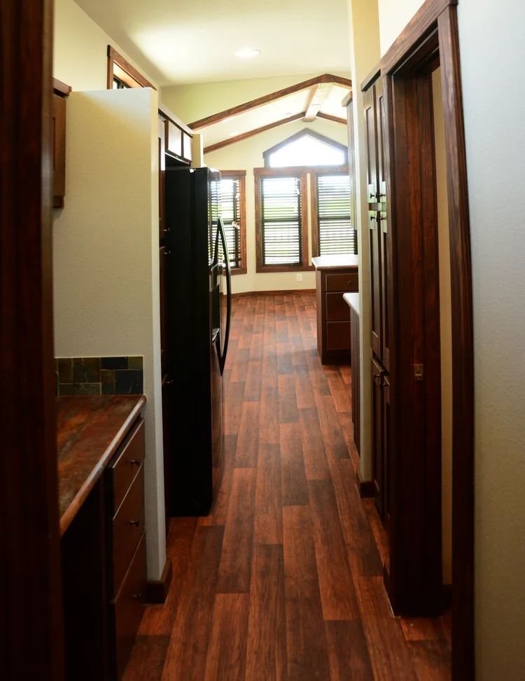 A narrow, well-lit kitchen with wooden floors, dark cabinets, and a high ceiling. Sunlight streams through large windows at the far end, adding warmth.