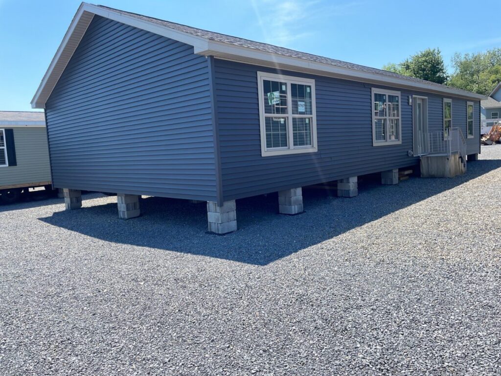 Blue manufactured home with white trim sits elevated on concrete blocks on a gravel lot. Sunlit sky adds a bright and serene tone.