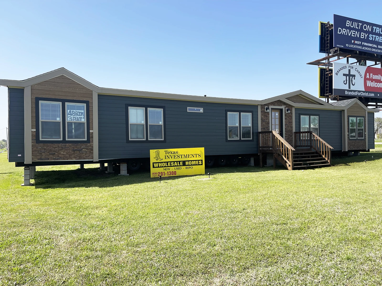A dark gray manufactured home with brown accents sits on a grassy field. A sign reads "Texas Investments Wholesale Homes." A billboard towers above.