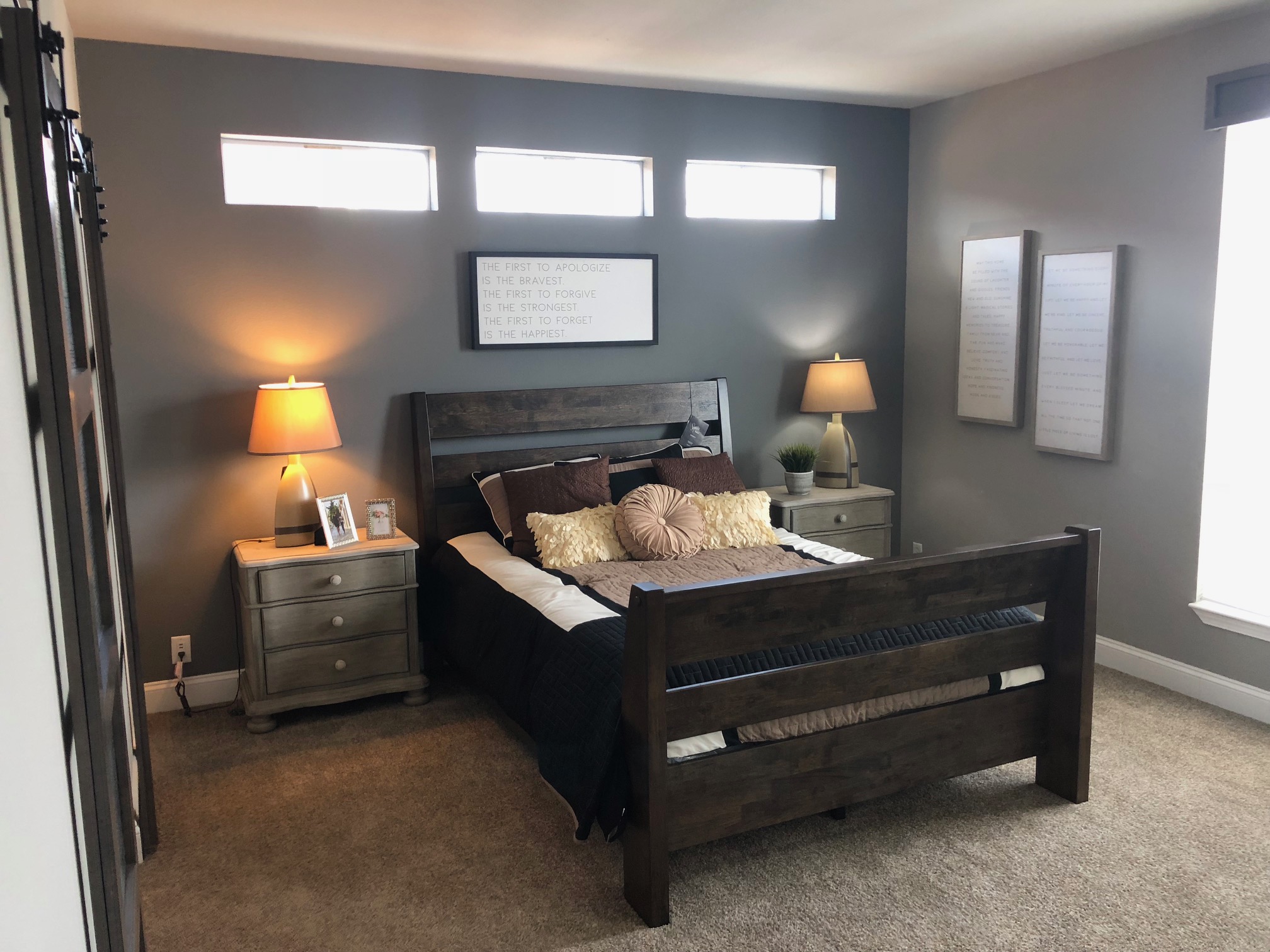 Cozy bedroom with a wooden bed, two bedside tables, and warm lamps. Walls are gray, adorned with framed quotes. Soft pillows and decor add warmth.