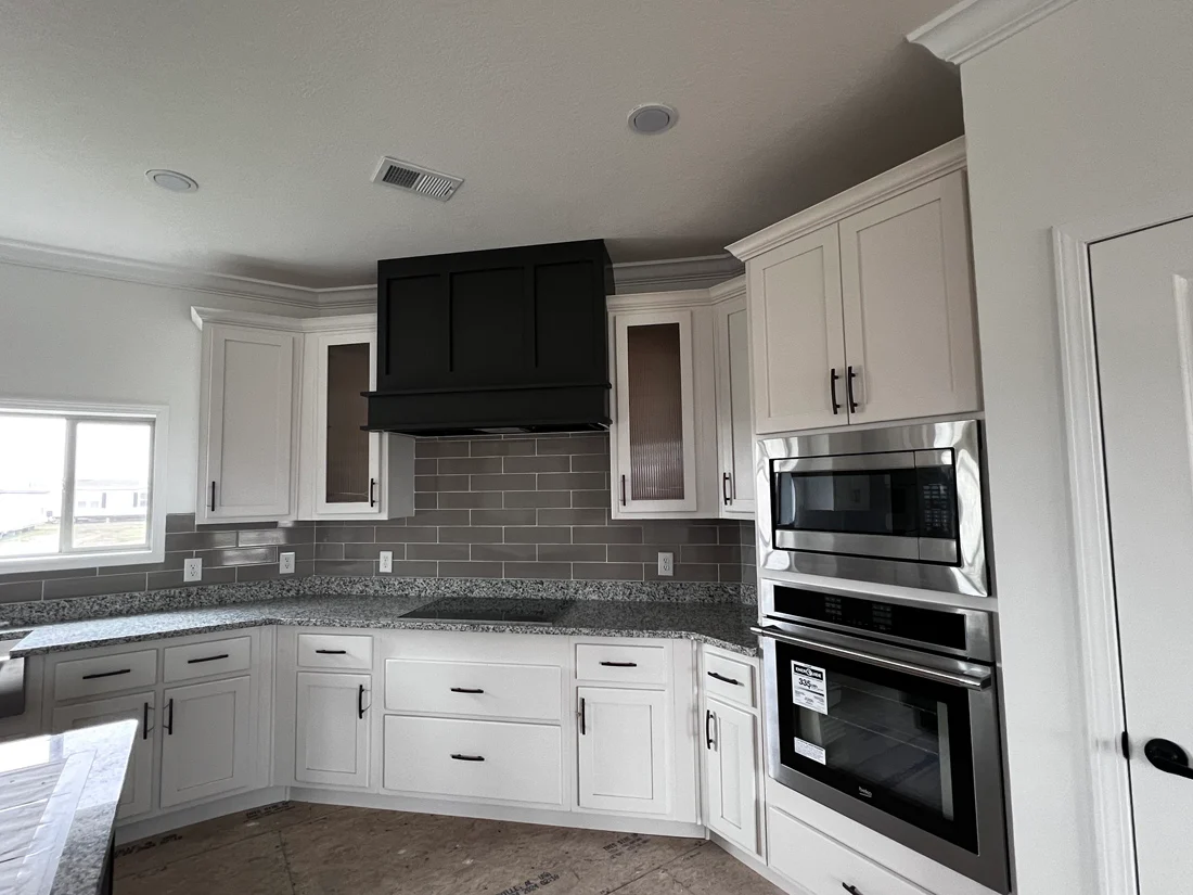 Modern kitchen with white cabinets, stainless steel double oven, black range hood, gray tile backsplash, and granite countertops. Bright and sleek design.