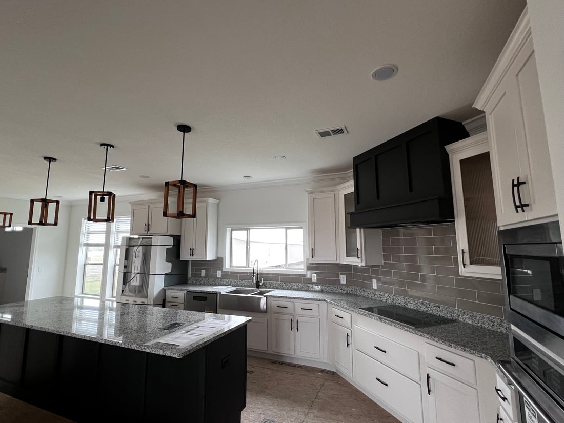 A modern kitchen with white cabinets, a black range hood, and gray tile backsplash. Granite countertops and island, pendant lights, and stainless steel appliances.