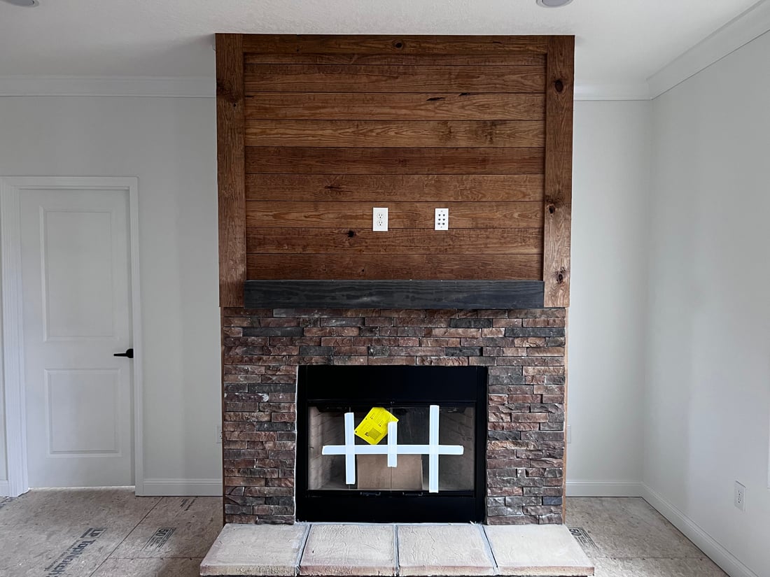 Rustic fireplace with wood paneling above and brick surround below. A wooden mantel sits above the stonework. The floor is unfinished plywood.