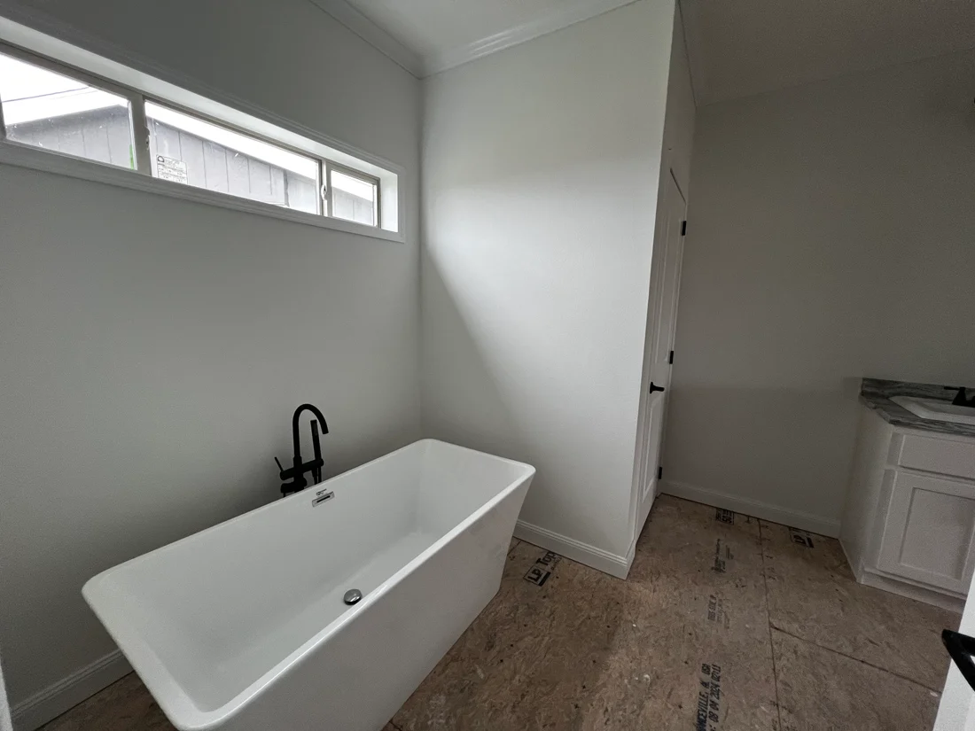 Minimalist bathroom with a white freestanding tub, black faucet, narrow window above, and a white sink cabinet. The space is under construction.