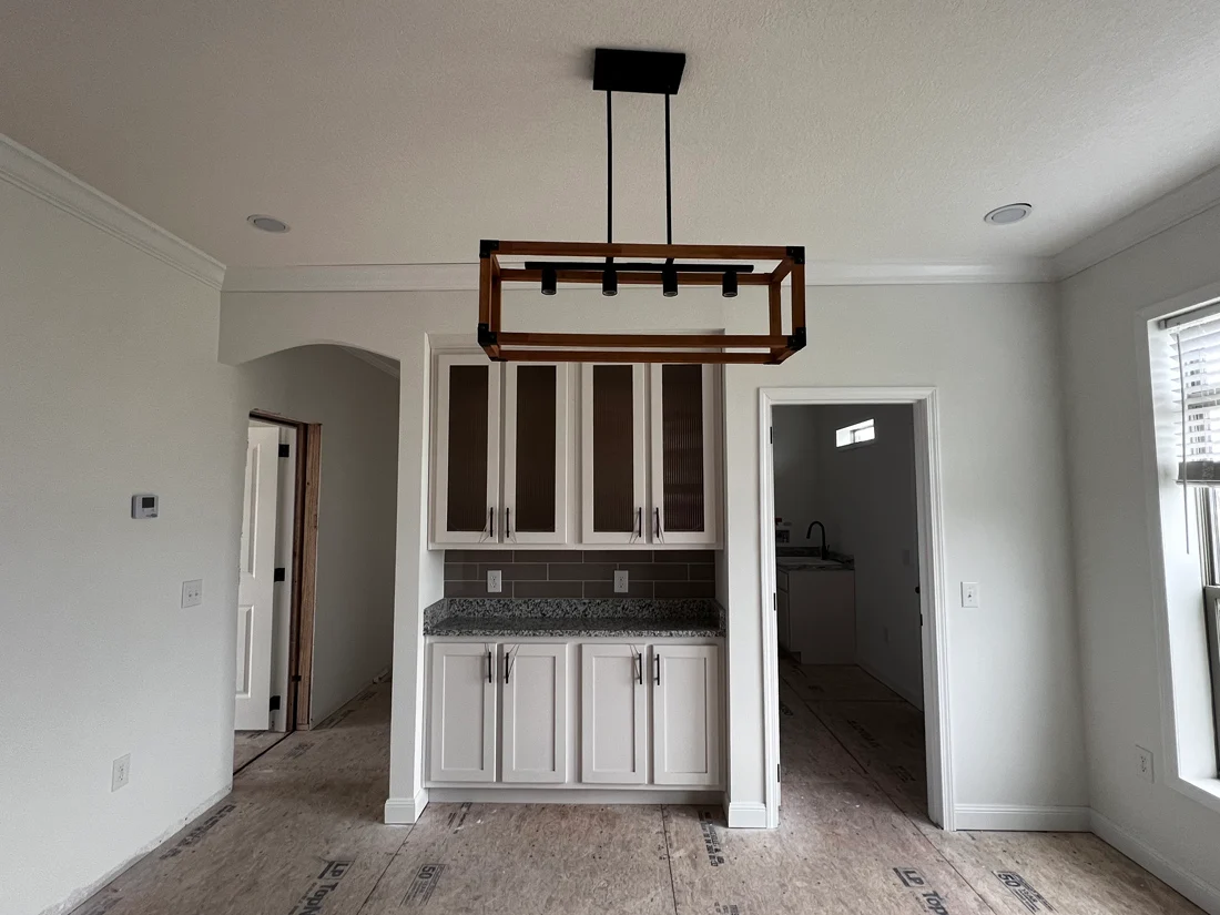 Modern kitchen with white cabinets, granite countertop, and a rectangular wooden light fixture above. Bright natural light from window on the right.